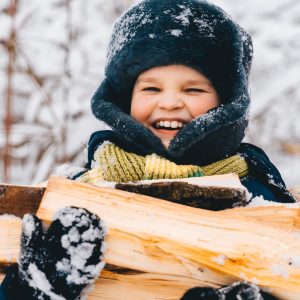 Candid portrait of smiling Boy at countryside backyard. Farm lifestyle. happy kid carries a heap of wood for making fire dressed in warm jacket, hat, gloves. child carries firewood to house in winter.